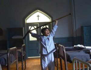 Scene from Indian Horse (movie); Indigenous child in a residential school happy and holding a hockey stick over head.